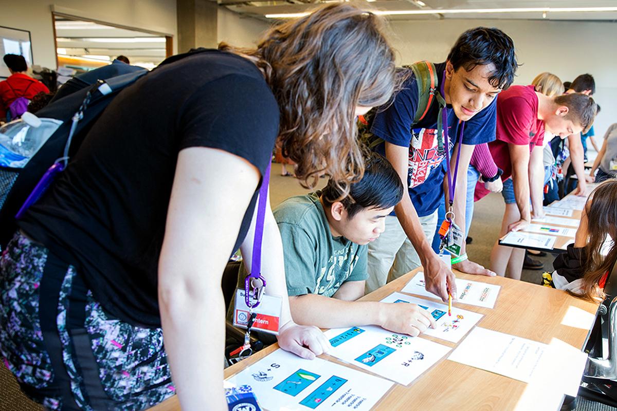 Three students of varying identities look at graphics on a table in a room full of other students at the Center for Neurotechnology.